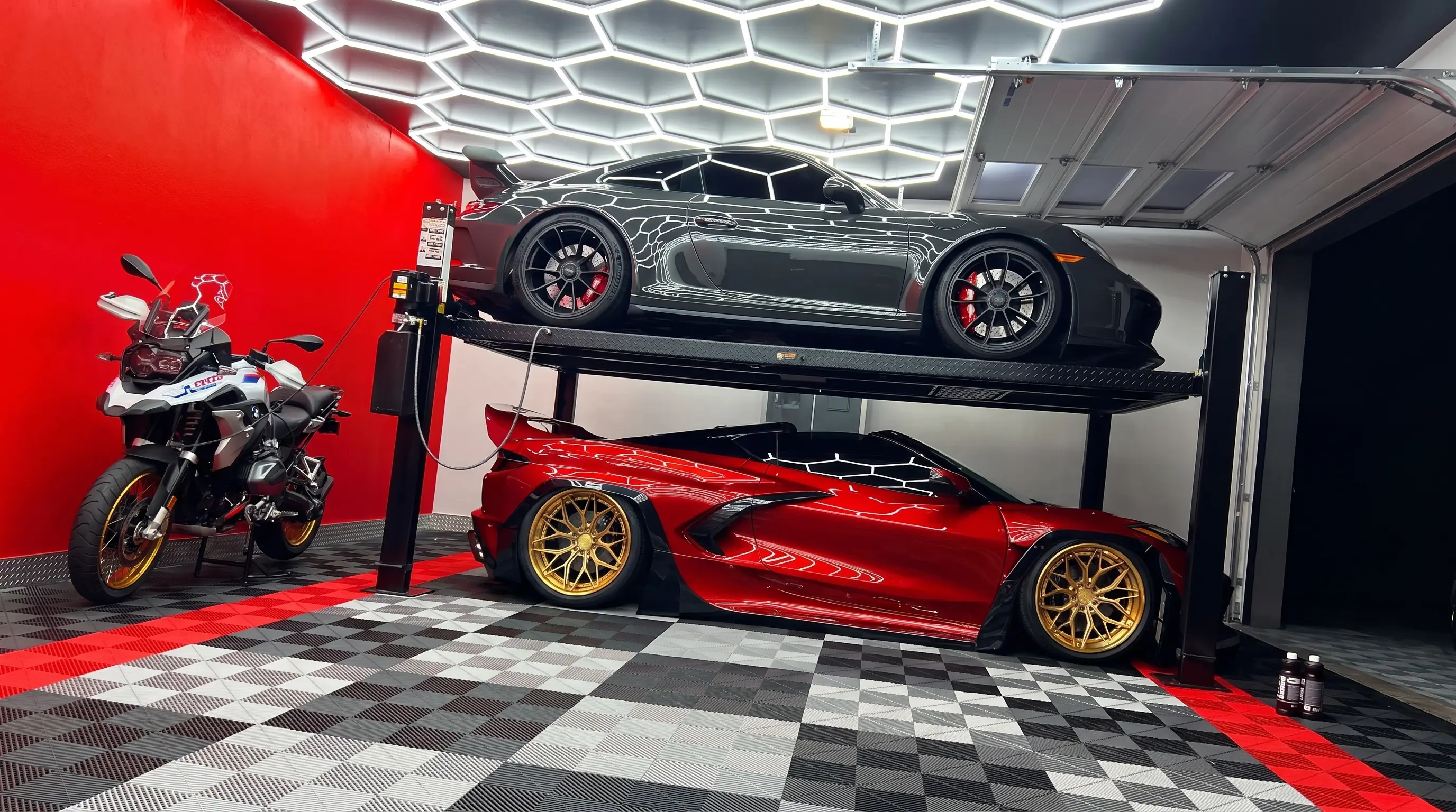 Two-post lift with a black coupe raised above a red Corvette, on a black, white and grey checkerboard tile floor with red border, under hexagon lighting
